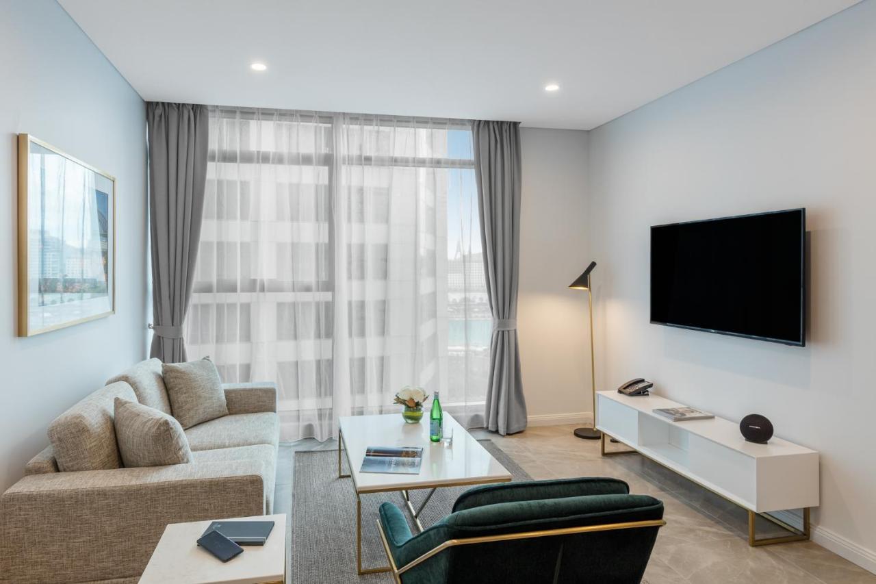 Meriton Suites Sussex Street, Sydney Ngoại thất bức ảnh The photo shows a modern living room space with a light, airy ambiance. It features a comfortable beige sofa with decorative cushions, a sleek coffee table placed in front, and a dark green armchair. There is a large window covered with sheer curtain