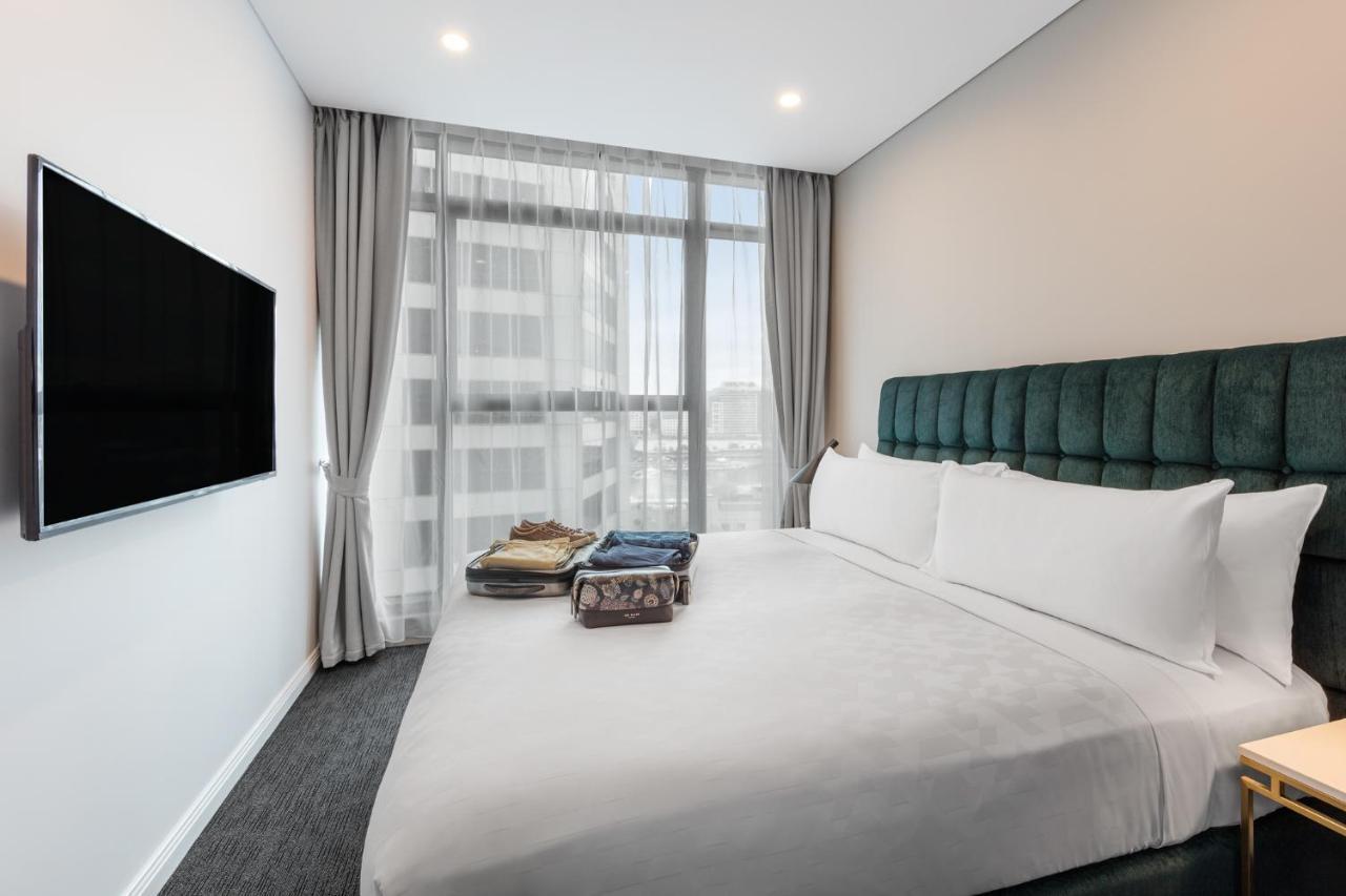 Meriton Suites Sussex Street, Sydney Ngoại thất bức ảnh The photo shows a modern hotel bedroom. The room features a large bed with a green upholstered headboard and crisp white linens. There are two pillows on the bed and a few folded items of clothing neatly placed on it. A flat-screen TV is mounted on t