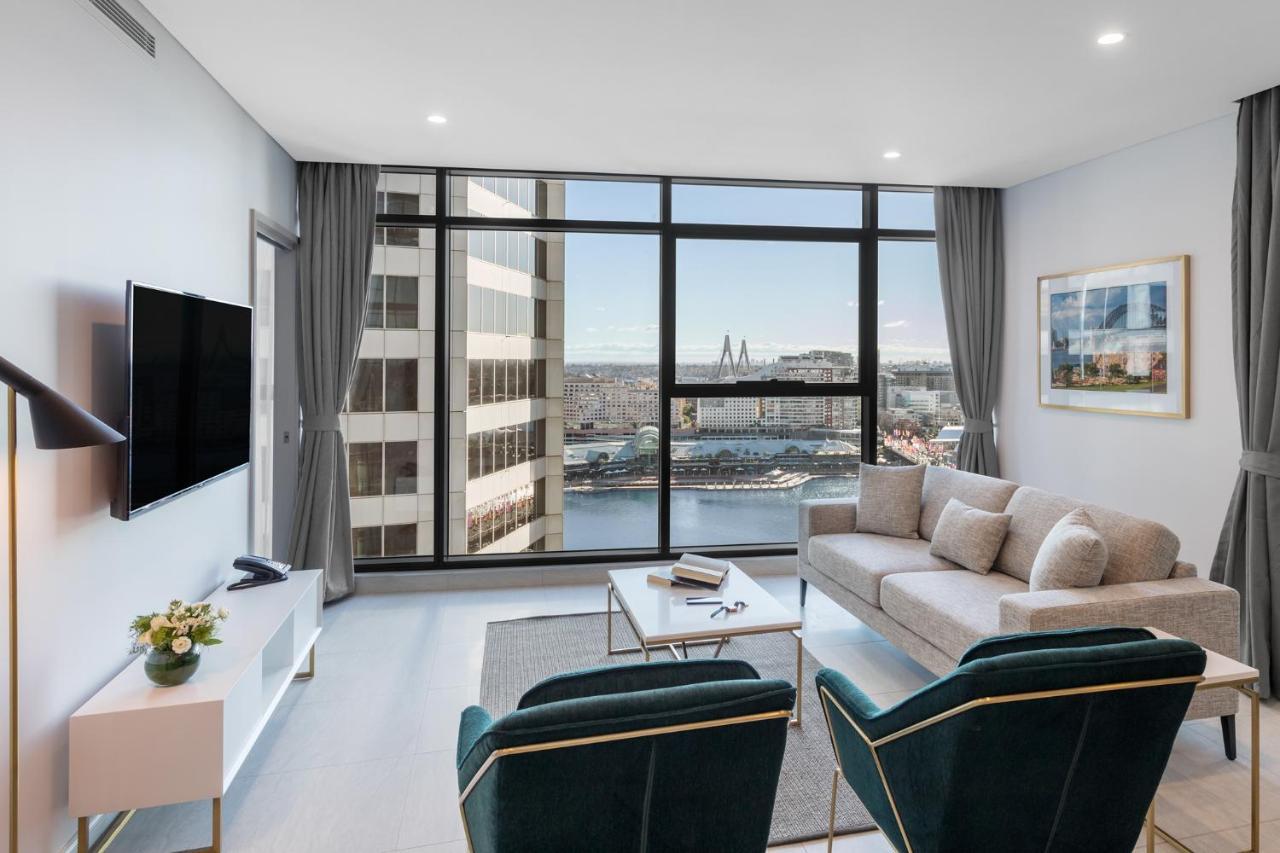Meriton Suites Sussex Street, Sydney Ngoại thất bức ảnh The photo shows a modern living room with large windows that offer a view of the cityscape and water. The interior features a light color palette with pale blue walls and soft lighting. There is a comfortable gray sofa and two green accent chairs, ar