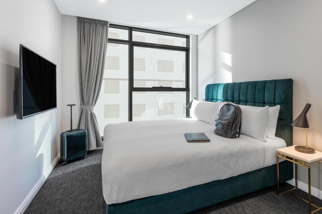 Meriton Suites Sussex Street, Sydney Ngoại thất bức ảnh The photo shows a modern hotel room featuring a comfortable bed with white bedding and a teal-colored headboard. There is a neatly placed black backpack on the bed, along with a small notebook or tablet. A television is mounted on the wall opposite t