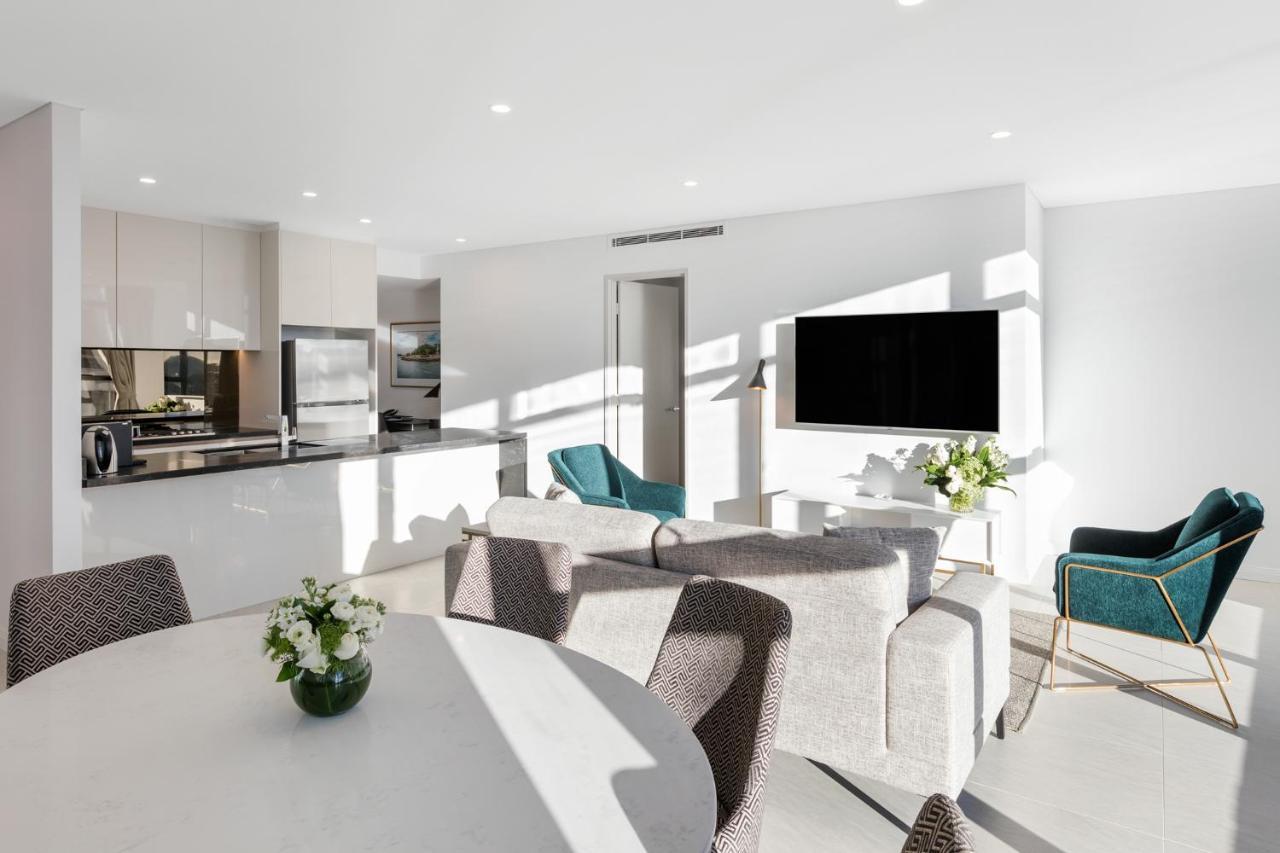 Meriton Suites Sussex Street, Sydney Ngoại thất bức ảnh The photo shows a modern, bright living space that combines a kitchen and a living area. The kitchen features sleek cabinets and stainless steel appliances. In the foreground, there is a round dining table with a vase of flowers on it. Seating option