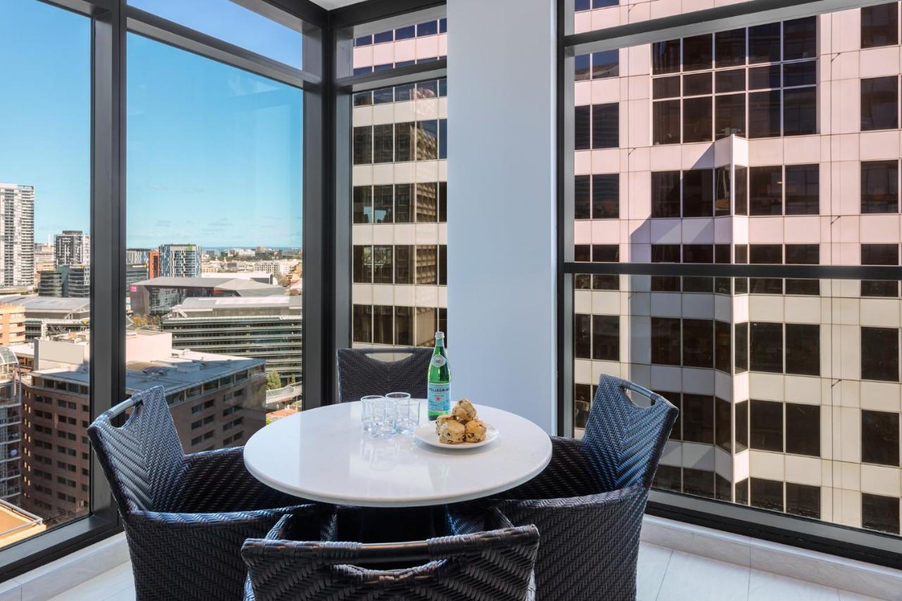 Meriton Suites Sussex Street, Sydney Ngoại thất bức ảnh The photo shows a modern dining space with a round white table set for four. There are four black woven chairs around the table. On the table, there's a bottle of sparkling water, two glasses, and a small plate with several cookies. The large windows