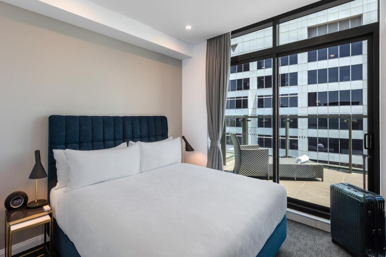 Meriton Suites Sussex Street, Sydney Ngoại thất bức ảnh The photo shows a modern hotel room with a large bed featuring a blue upholstered headboard and white linens. Natural light enters through a large window, revealing a view of a building exterior, likely part of the hotel or surrounding area. The room