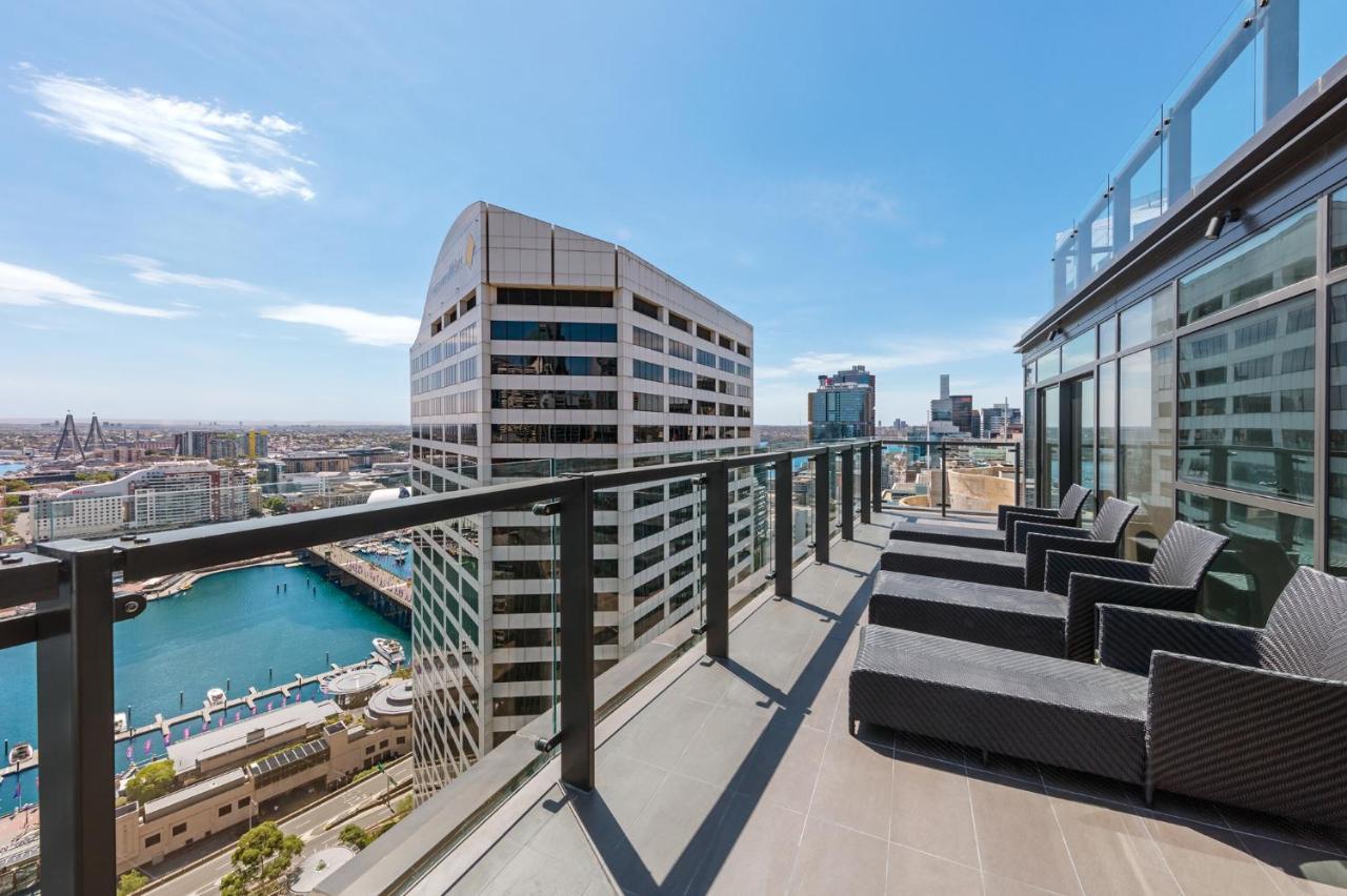 Meriton Suites Sussex Street, Sydney Ngoại thất bức ảnh The photo shows a balcony or terrace with a panoramic view of a city skyline. The balcony features modern outdoor furniture, including sleek lounge chairs. In the background, you can see a waterfront area with buildings and a harbor, along with a cle