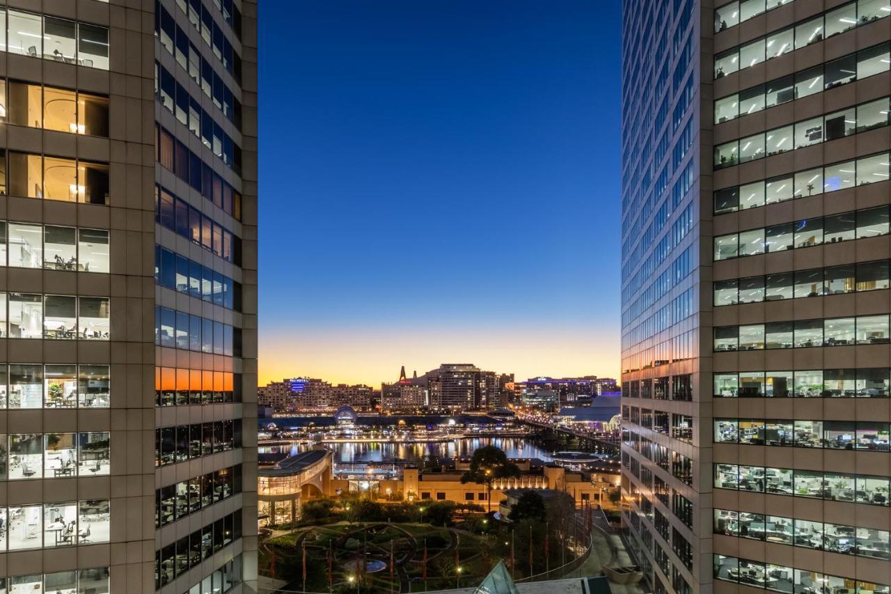 Meriton Suites Sussex Street, Sydney Ngoại thất bức ảnh The photo shows a view between two tall buildings, likely office towers, during twilight. On one side, there are glass windows reflecting the evening sky that transitions from blue to orange near the horizon. In the background, a cityscape is visible