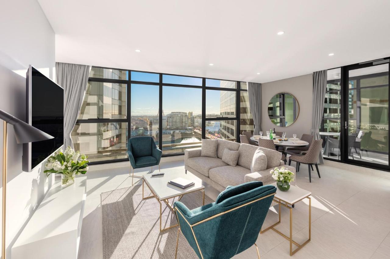 Meriton Suites Sussex Street, Sydney Ngoại thất bức ảnh The photo shows a modern and stylish living space. It features a spacious room with large windows that provide a bright view of the cityscape. The furniture includes a light gray sofa, two teal chairs, and a coffee table. There is a flat-screen TV mo
