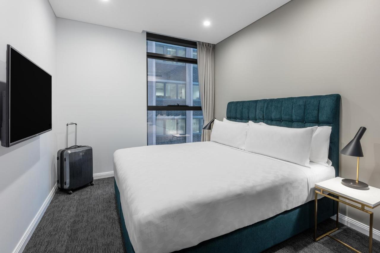 Meriton Suites Sussex Street, Sydney Ngoại thất bức ảnh The photo shows a modern hotel room with a clean and minimalist design. There is a large, neatly made bed with white bedding and a teal-colored headboard. On one side of the bed, there is a small nightstand with a lamp. A sleek television is mounted 