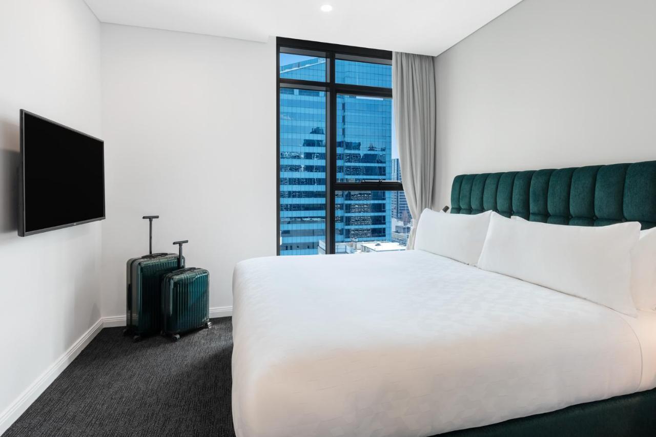 Meriton Suites Sussex Street, Sydney Ngoại thất bức ảnh The photo shows a modern hotel room featuring a large bed with white bedding and a green upholstered headboard. There are two green suitcases placed beside the bed. A flat-screen TV is mounted on the wall opposite the bed. The room has a large window