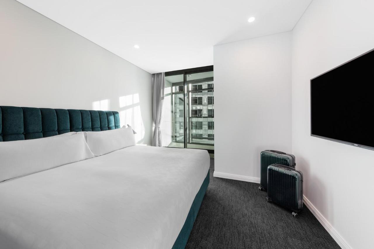 Meriton Suites Sussex Street, Sydney Ngoại thất bức ảnh The photo shows a modern hotel room featuring a large bed with a headboard in a teal color. The bedding is white and appears neatly arranged. There is a large window allowing natural light to enter, offering a view of an outdoor area. A flat-screen T