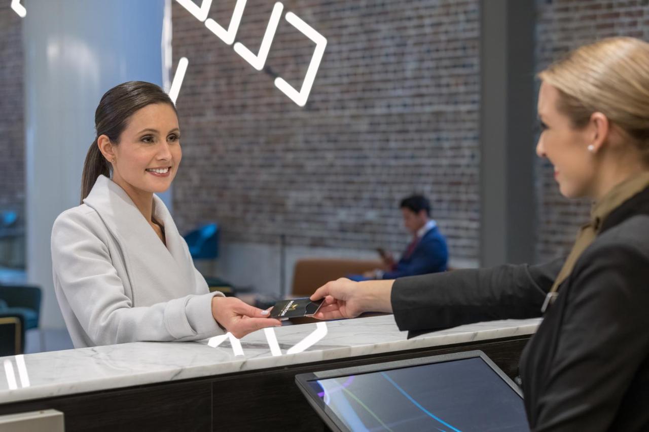 Meriton Suites Sussex Street, Sydney Ngoại thất bức ảnh The image depicts a scene at a hotel reception desk. A woman with long hair, dressed in a white outfit, is smiling as she hands her card to a receptionist, who is in a black uniform. The reception area features a modern design, and there is a person 