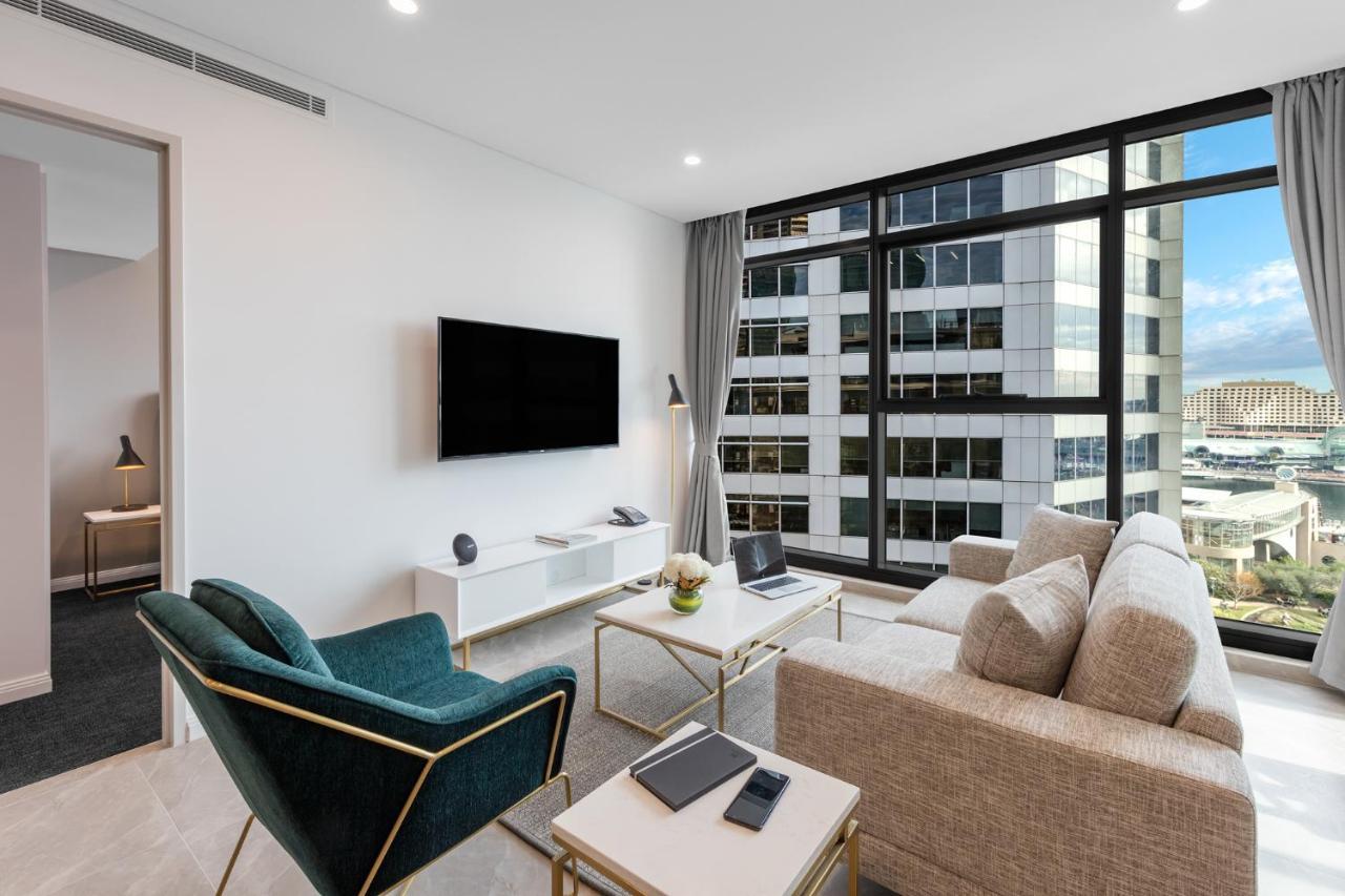 Meriton Suites Sussex Street, Sydney Ngoại thất bức ảnh The photo shows a modern living room with large windows that let in natural light, providing a view of a cityscape outside. The interior features a comfortable light-colored sofa and a stylish dark green armchair. A sleek TV is mounted on the wall op