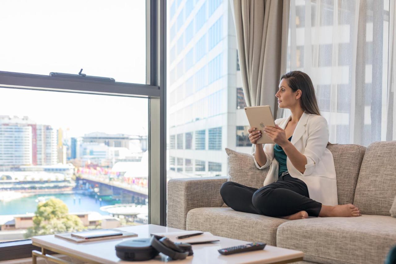 Meriton Suites Sussex Street, Sydney Ngoại thất bức ảnh The image portrays a woman sitting cross-legged on a light-colored sofa, engaged with a tablet in her hands. She is dressed in a white blazer and dark pants, appearing relaxed and focused. The setting features large windows that provide a view of a w