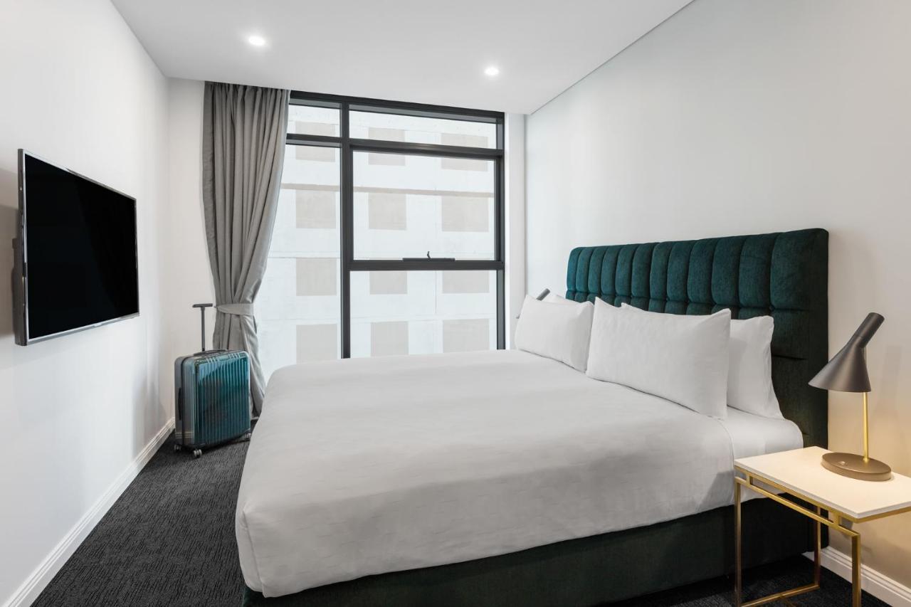 Meriton Suites Sussex Street, Sydney Ngoại thất bức ảnh The photo shows a modern hotel room. It features a large bed with a dark green tufted headboard and multiple white pillows. There's a nightstand with a lamp beside the bed. A sleek television is mounted on the wall opposite the bed, and a suitcase is