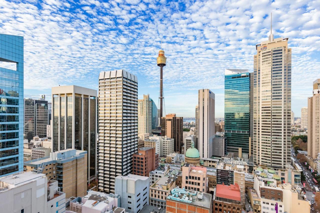 Meriton Suites Sussex Street, Sydney Ngoại thất bức ảnh The photo shows a panoramic view of a city skyline dominated by tall skyscrapers. In the center, there is a notable tower with a distinctive golden structure at its top, which is likely a landmark of the city. The buildings have a mix of modern glass