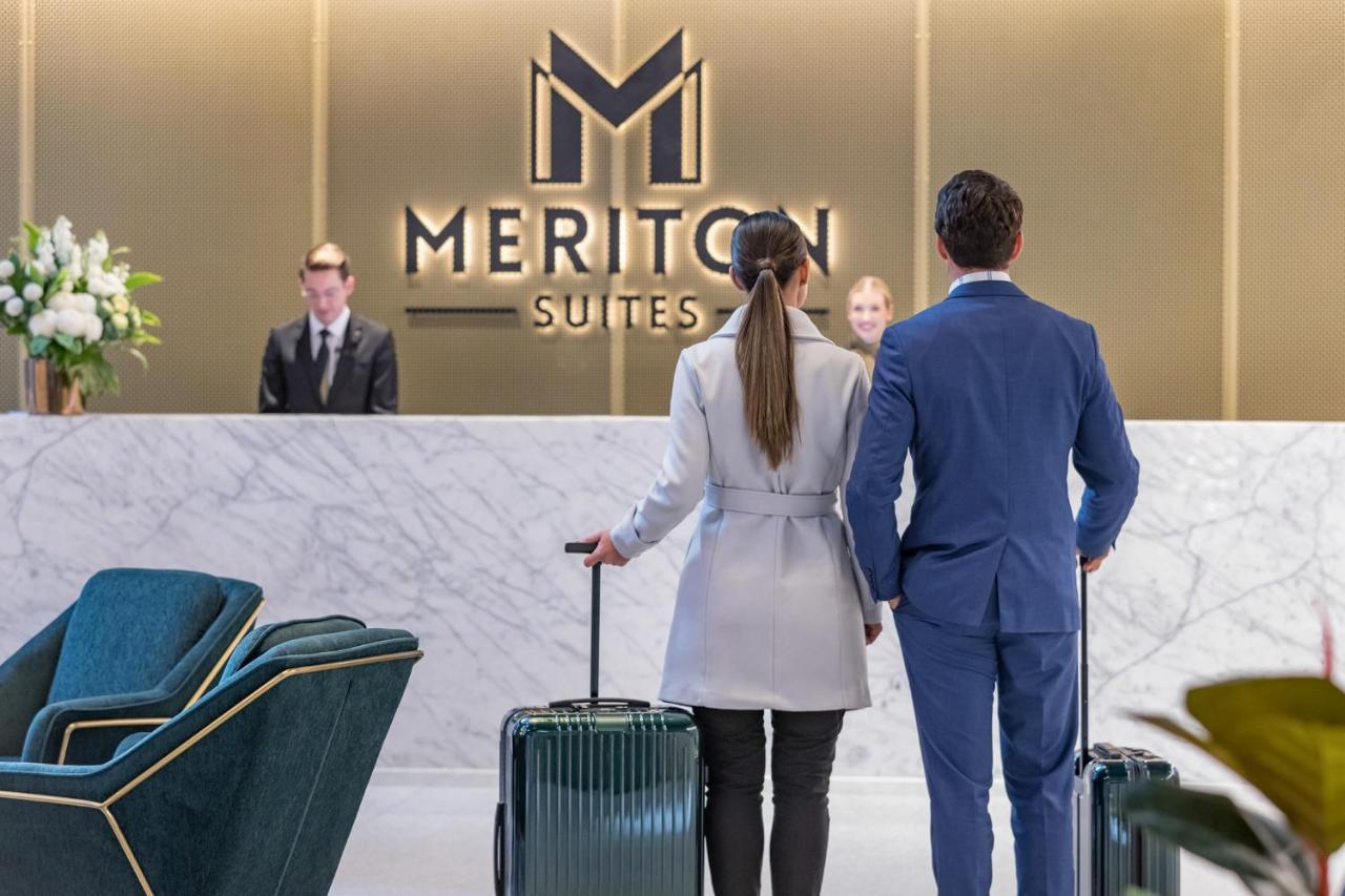 Meriton Suites Sussex Street, Sydney Ngoại thất bức ảnh The photo shows a couple standing at a reception area in a hotel named "Meriton Suites." They are both facing the reception desk, where staff members are visible. The man is dressed in a suit, and the woman is wearing a light-colored coat. They each 