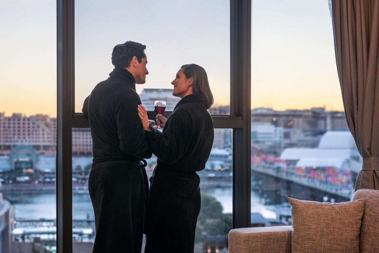 Meriton Suites Sussex Street, Sydney Ngoại thất bức ảnh The photo depicts a couple standing closely together by a large window. They are wearing dark bathrobes and holding drinks, gazing into each other’s eyes. The background features a scenic view, likely of a cityscape at sunset, with soft light illumin