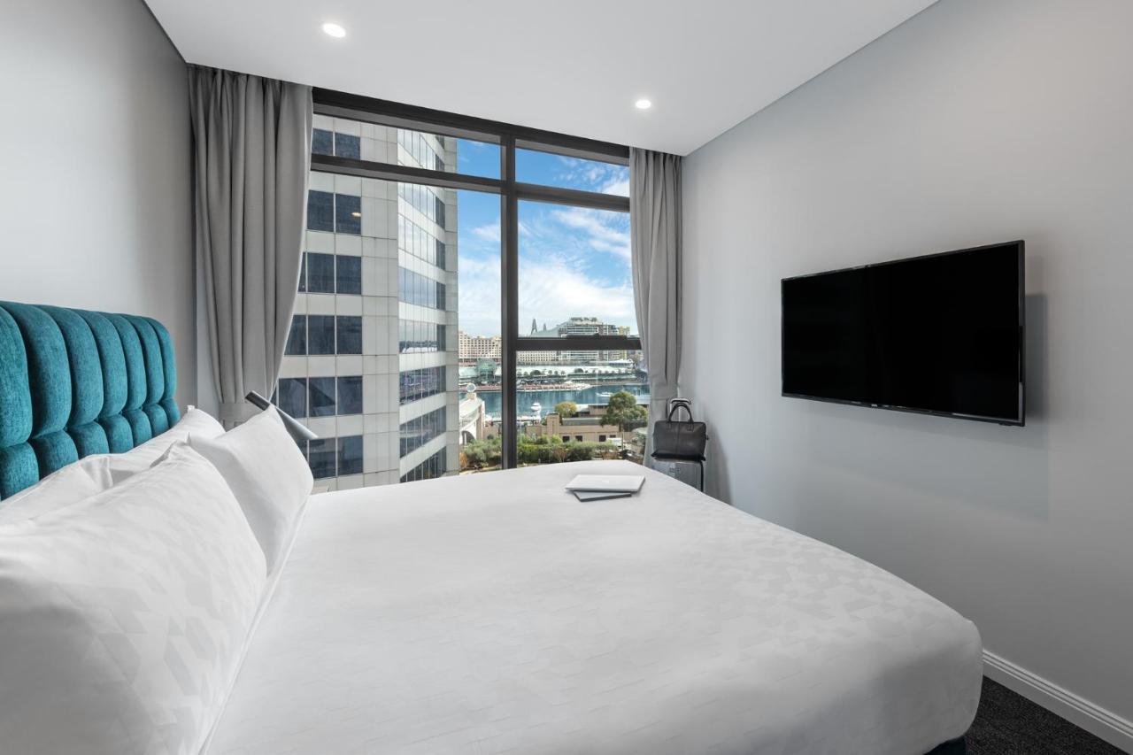 Meriton Suites Sussex Street, Sydney Ngoại thất bức ảnh The photo shows a modern hotel room with a large bed featuring a white comforter and pillows. There is a blue headboard that adds a pop of color to the decor. A large window provides a view of the outside, which includes buildings and a glimpse of wa