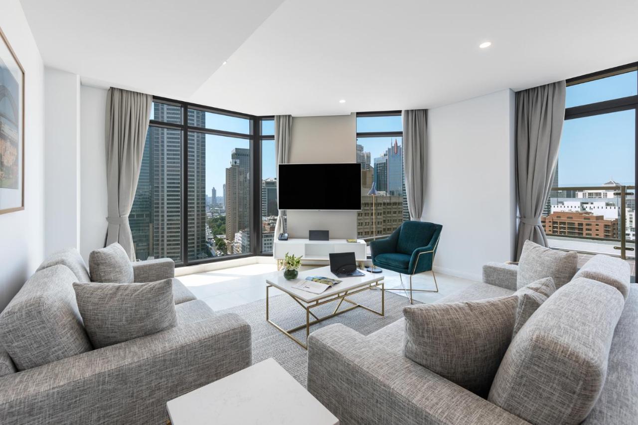 Meriton Suites Sussex Street, Sydney Ngoại thất bức ảnh The photo showcases a modern living room with large windows offering a view of a city skyline. The room features a light color palette with gray sofas arranged around a coffee table. A flat-screen TV is mounted on the wall, and there is a green armch