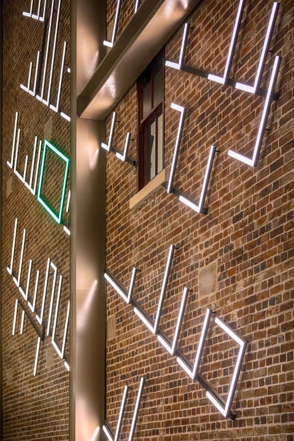 Meriton Suites Sussex Street, Sydney Ngoại thất bức ảnh The photo shows a brick wall featuring a design made up of linear light fixtures. These light strips create geometric shapes and patterns, with some sections illuminated in white and one section highlighted in green. The combination of the brick text