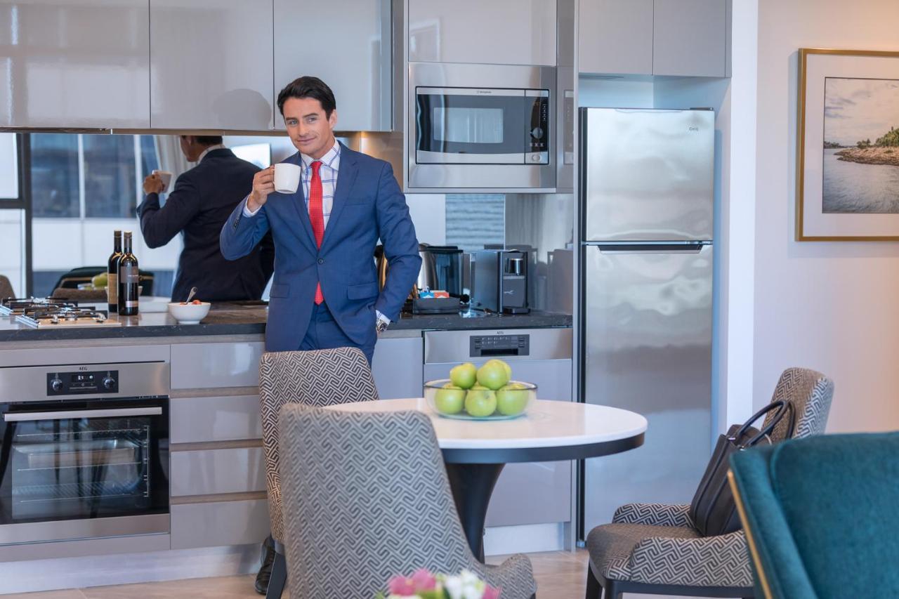 Meriton Suites Sussex Street, Sydney Ngoại thất bức ảnh The photo depicts a well-dressed man in a suit standing in a modern kitchen. He is holding a cup, possibly of coffee, and appears to be looking towards the camera. The kitchen features contemporary appliances, including a microwave and refrigerator. 