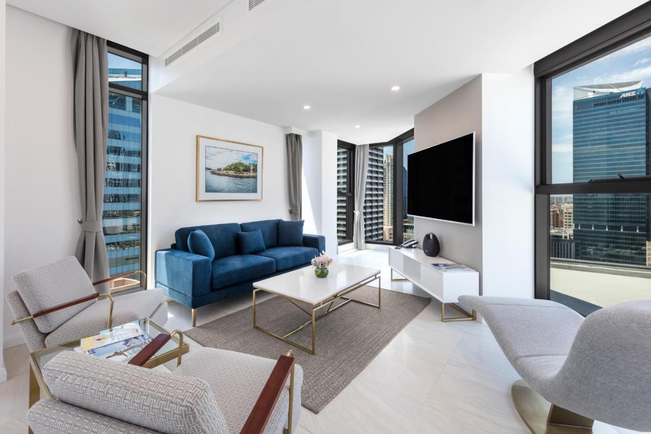 Meriton Suites Sussex Street, Sydney Ngoại thất bức ảnh The photo shows a modern living room featuring large windows that allow plenty of natural light. The room is elegantly designed with a blue sofa, a light-colored coffee table, and two armchairs. There’s a flat-screen TV mounted on the wall, and a pie